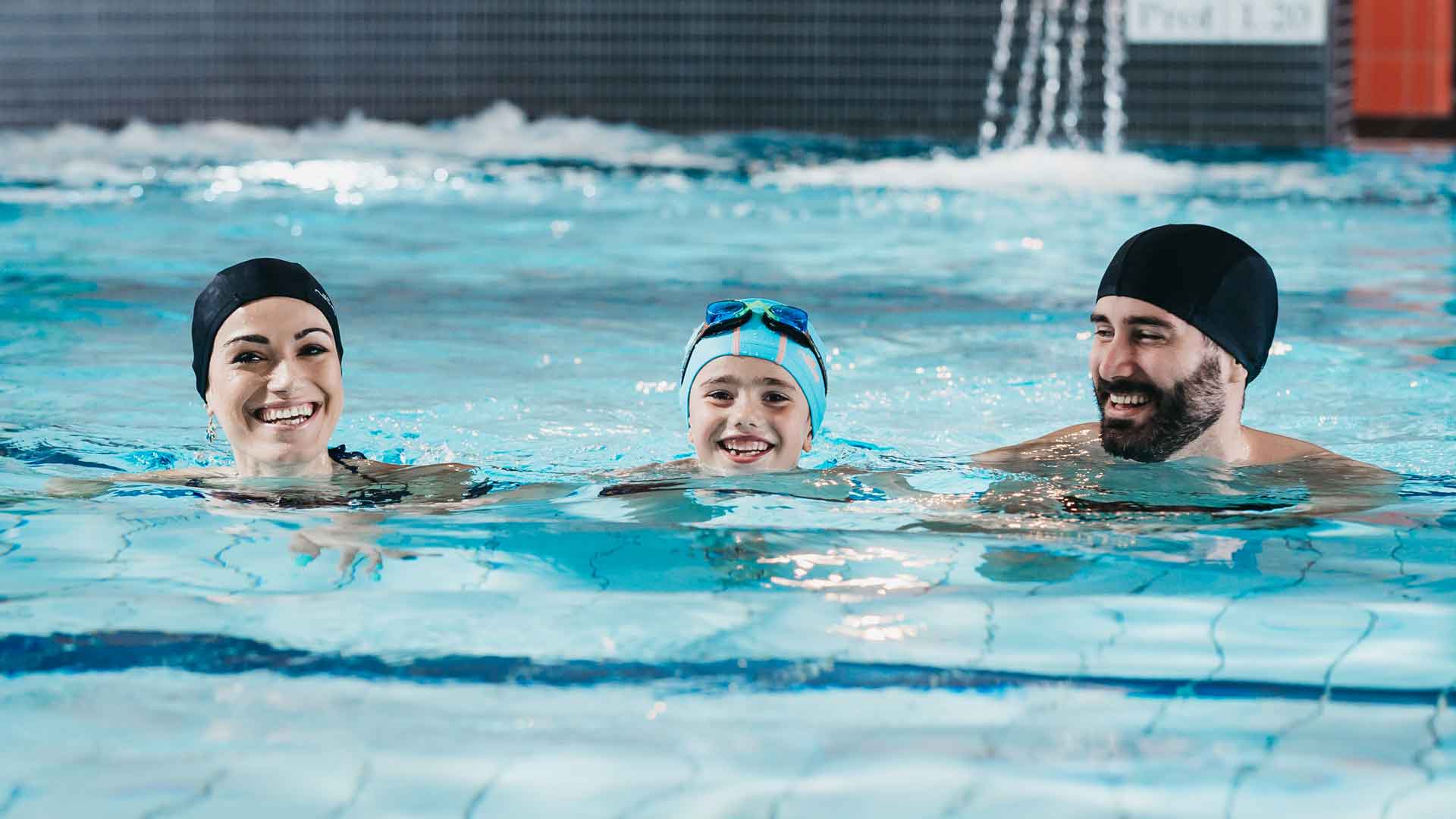 Un enfant et deux adultes en maillot de bain avec un bonnet de bain, sourient face à l'objectif, dans la piscine.